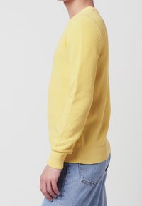 Gelbes, gestricktes Sweatshirt mit einem strukturierten Muster, langen Ärmeln und gerippten Bündchen, kombiniert mit hellblauen Jeans.