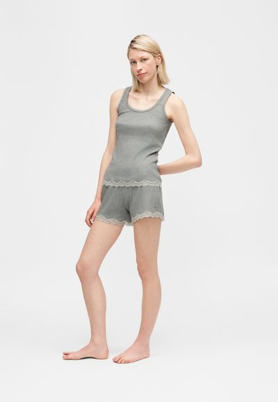Szőke nő mezítláb áll, szürke bordázott ujjatlan felsőt és hozzáillő rövidnadrágot visel, amely csipkés szegéllyel díszített, fehér háttér előtt.