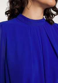 Royal blue blouse with high neck, soft fabric, and pleated design at the front. Transparent sleeves add delicate texture.