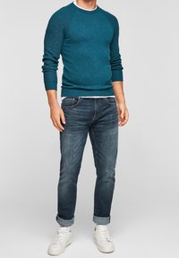 Maglione color turchese lavorato a maglia con texture a coste, abbinato a jeans di denim scuro e sneakers bianche. Il maglione presenta maniche raglan e scollo tondo.