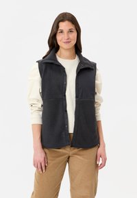 Schwarze Fleece-Weste mit einem Reißverschlusskragen, Druckknopf-Front und kontrastierenden Stoffpaneelen. Präsentiert über einem hellcremefarbenen Pullover und beigen Hosen.