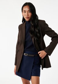 Bruin op maat gemaakte blazer over een marineblauwe coltrui, gecombineerd met een marineblauwe rok met een riem. Soepele stof met een gestructureerde silhoeet.