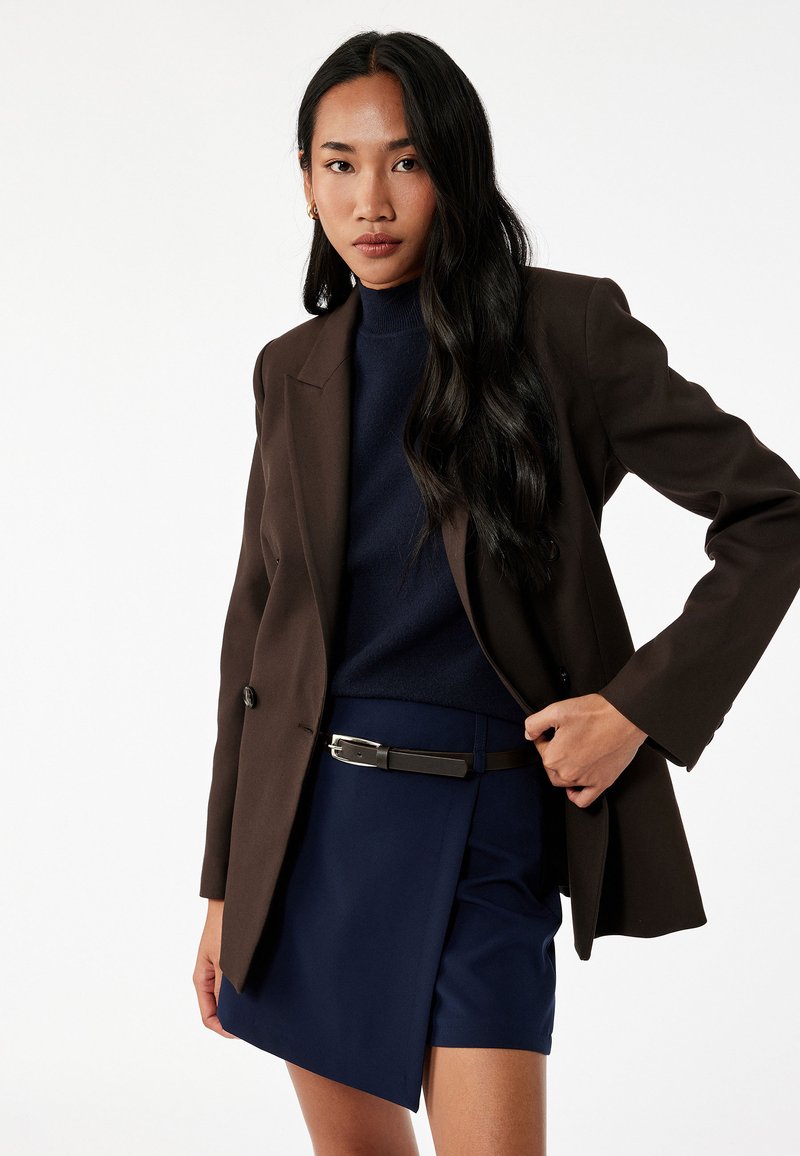 Bruin op maat gemaakte blazer over een marineblauwe coltrui, gecombineerd met een marineblauwe rok met een riem. Soepele stof met een gestructureerde silhoeet.