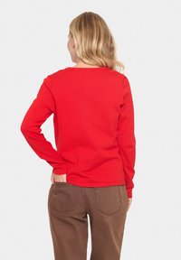 Haut rouge à manches longues avec un col ras du cou, design ajusté, texture lisse. Porté avec un pantalon marron taille haute ; vue de dos visible.