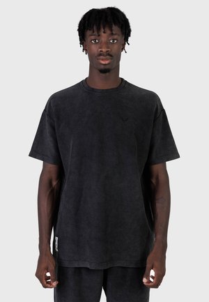 Junger Mann mit kurzen Dreadlocks, der ein locker sitzendes schwarzes Cord-T-Shirt und passende Hose trägt, vor einem schlichten Hintergrund stehend.