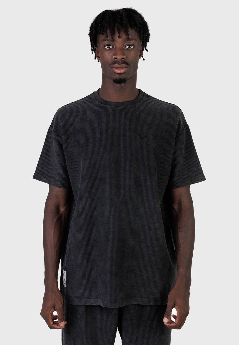 Joven con dreadlocks cortos vistiendo una camiseta de pana negra holgada y pantalones a juego, de pie frente a un fondo liso.