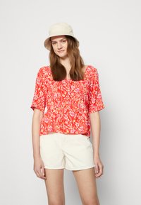 Blusa floral vermelha com mangas curtas, decote quadrado e detalhes franzidos, emparelhada com calções creme e um chapéu estilo balde de cor clara.