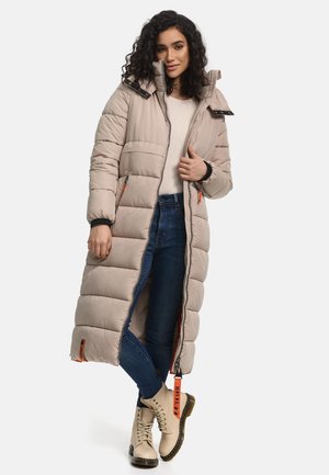 Long manteau en duvet beige avec fermeture éclair à l'avant, col haut et poignets élastiques. Doté de poches latérales avec des accents orange. Associé à un jean bleu et des bottes beiges.