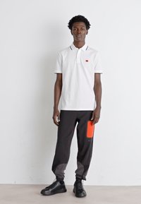 Polo bianco con un piccolo logo rosso, abbinato a pantaloni neri con una tasca arancione brillante. Scarpe nere completano il look.