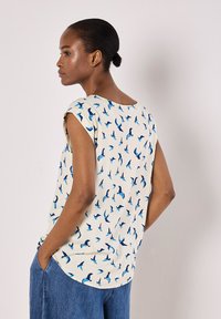 Haut sans manches crème avec un motif d'oiseaux bleus, décolleté arrondi et coupe décontractée. La vue de dos montre un ourlet légèrement incurvé et un tissu léger.