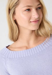 Mujer joven con cabello rubio que lleva un suéter de punto ligero en color púrpura, mirando ligeramente de lado con una sonrisa sutil.