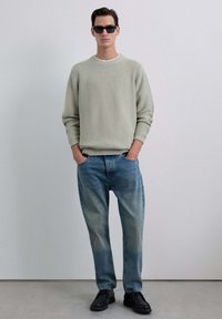 Pull en maille gris clair avec un col rond, assorti à un jean bleu délavé et des chaussures noires. Texture lisse et coupe décontractée.