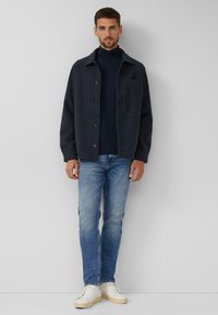 Marineblaue Jacke mit Knopfleiste und einer Brusttasche, getragen über einem Rollkragenpullover, kombiniert mit hellen Jeans und weißen Sneakern.