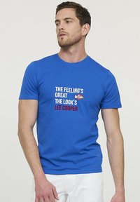 T-shirt en coton bleu à manches courtes avec texte imprimé en blanc et rouge, présentant les phrases "C'EST SUPER" et "LE LOOK EST LEE COOPER."