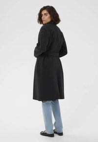 Manteau en mélange de laine noir avec une ceinture, un col haut et des manches longues, associé à un jean bleu clair et des mocassins noirs.