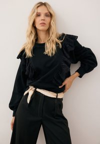 Czarny sweatshirt z falbanami, z haftowanym wzorem, połączony z czarnymi spodniami o wysokim stanie i kremowym paskiem.