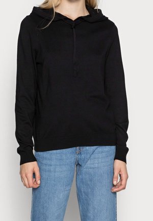 Personne portant un sweat à capuche noir uni à manches longues avec cordons et un jean en denim bleu clair sur un fond uni.
