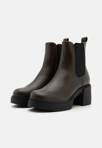 Bottines Chelsea en cuir marron avec un large talon noir et des panneaux latéraux élastiques. Texture lisse avec des détails de couture минимaux.