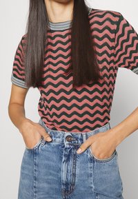 Dame kortærmet top med et bølget rødt og sort mønster, med en stribet halsudskæring. Parret med højtaljede denimshorts.