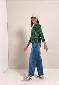 Haut à manches longues vert à motifs, jean en denim bleu ample et baskets grises avec des accents verts. Modèle portant une casquette claire, souriant.