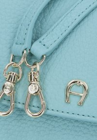 Hellblaue Lederhandtasche mit strukturierter Oberfläche, ausgestattet mit zwei Clips in Goldoptik und einem geprägten Logo-Akzent. Kurze, flache Griffe.