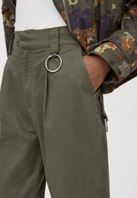 Pantalon kaki en tissu lisse avec un pli sur le devant, de grandes poches latérales et un accent en forme de cercle argenté à la taille.