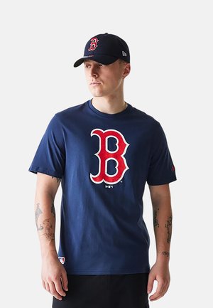 Mann trägt eine marineblaue Boston Red Sox Kappe und ein passendes T-Shirt mit großem rotem "B"-Logo, blickt nach links vor einem schlichten Hintergrund.