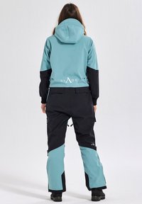 A dual-tone ski suit with a light teal upper and black lower sections. Features a hood, zippers, and logo detail on the back.