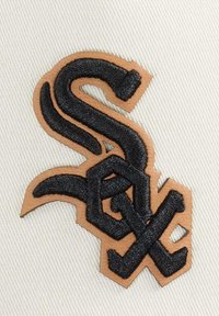 Schwarzes gesticktes "Sox"-Logo auf beige-farbener Material, mit ineinander verschlungenen Buchstaben und einer strukturierten Oberfläche auf einem hellen Hintergrund.