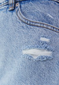 Vaaleansininen denim, jossa on rispaantunut, kulunut kohta; siinä on hienovaraisia sävyeroja ja näkyvää ompelua. Teksturoitu kangas näyttää kuluneelta.