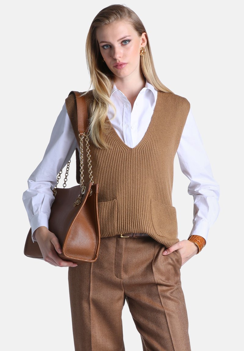 Femme portant une chemise blanche, un gilet en maille marron et un pantalon marron, tenant un grand sac à main en cuir marron avec des anses en chaîne dorée.