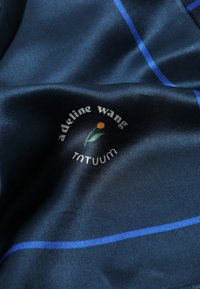 Tessuto satin blu navy con linee azzurre, caratterizzato da un logo con un fiore e il testo: "adeline wang TNTUUM." Texture liscia e finitura riflettente.