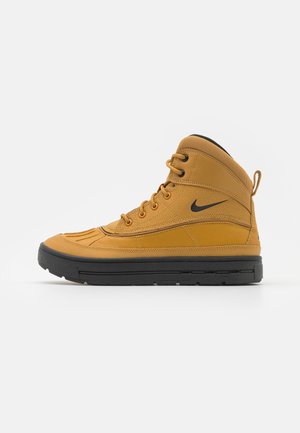 Høj top, sennepsgul Nike-støvle med sort sål, snørelukning foran, trækflig ved hælen og sort Nike Swoosh-logo på siden.