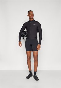 Black cycling jersey with a front zipper and long sleeves, paired with black shorts. The model holds a black cycling helmet in one hand.
