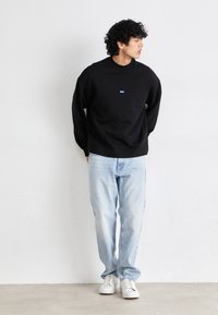 Zwarte sweatshirt met ronde hals, blauwe logo op de borst, gecombineerd met lichtblauwe denim jeans en witte sneakers. Staand tegen een effen achtergrond.