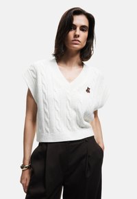Hvid kabelstrikket sweater med v-udskæring og korte ærmer, der har en lille farvet broderet bjørn detalje på venstre bryst.