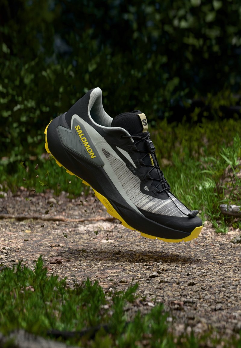 Scarpa da trail running con tomaia nera e grigia, dettagli gialli, suola texturizzata e intersuola ammortizzata per aderenza e comfort in ambienti esterni.