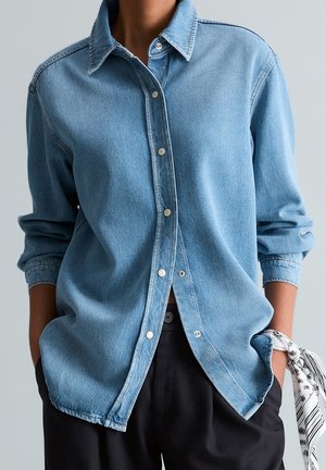 Persoon draagt een lichtblauw denim overhemd met knopen en opgerolde mouwen, zwarte broek en een witte sjaal met patroon, geknoopt om de pols.
