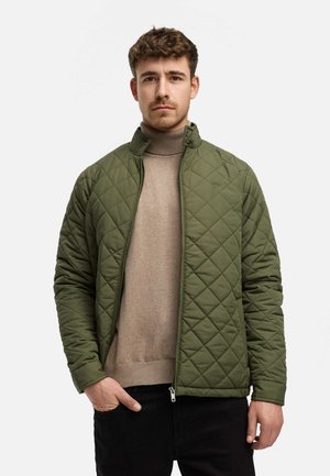 Quilted green jacket with a high collar and diagonal stitching, paired with a beige turtleneck and black pants. Zipper closure and side pockets.