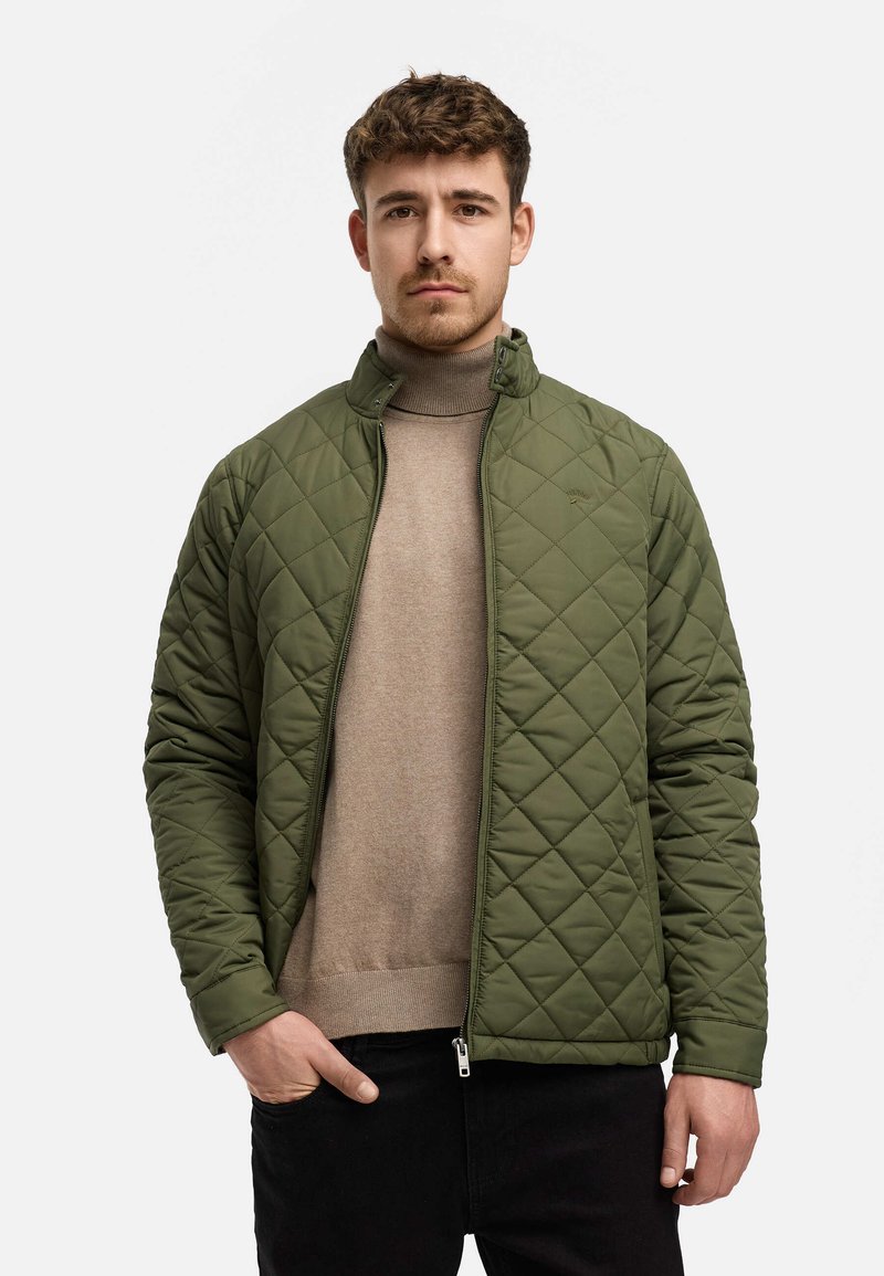 Quiltet grøn jakke med høj krave og diagonal syning, parret med en beige turtleneck og sorte bukser. Lynlåslukning og sidelommer.