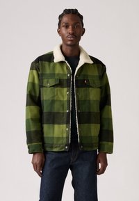 Chaqueta de cuadros verdes con acentos negros y un cuello de forro polar blanco. Presenta botones a presión y bolsillos frontales. Llevada sobre una camisa oscura.