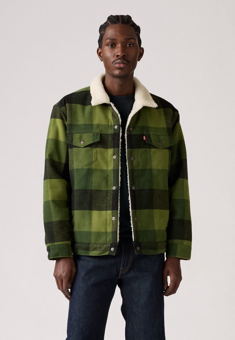 Chaqueta de cuadros verdes con acentos negros y un cuello de forro polar blanco. Presenta botones a presión y bolsillos frontales. Llevada sobre una camisa oscura.