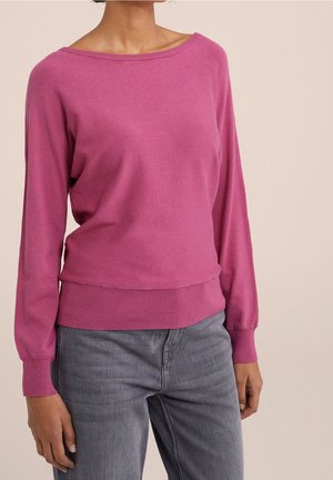 Femme portant un pull à manches longues rose vif avec des poignets et un ourlet côtelés, associé à un jean gris délavé sur un fond neutre.