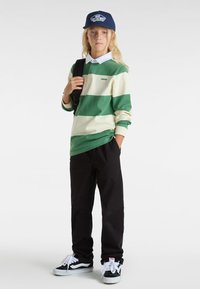 Gestreepte groene en roomkleurige lange mouwen shirt, zwarte broek, marineblauwe pet en zwarte sneakers met witte accenten, staande met een rugzak.