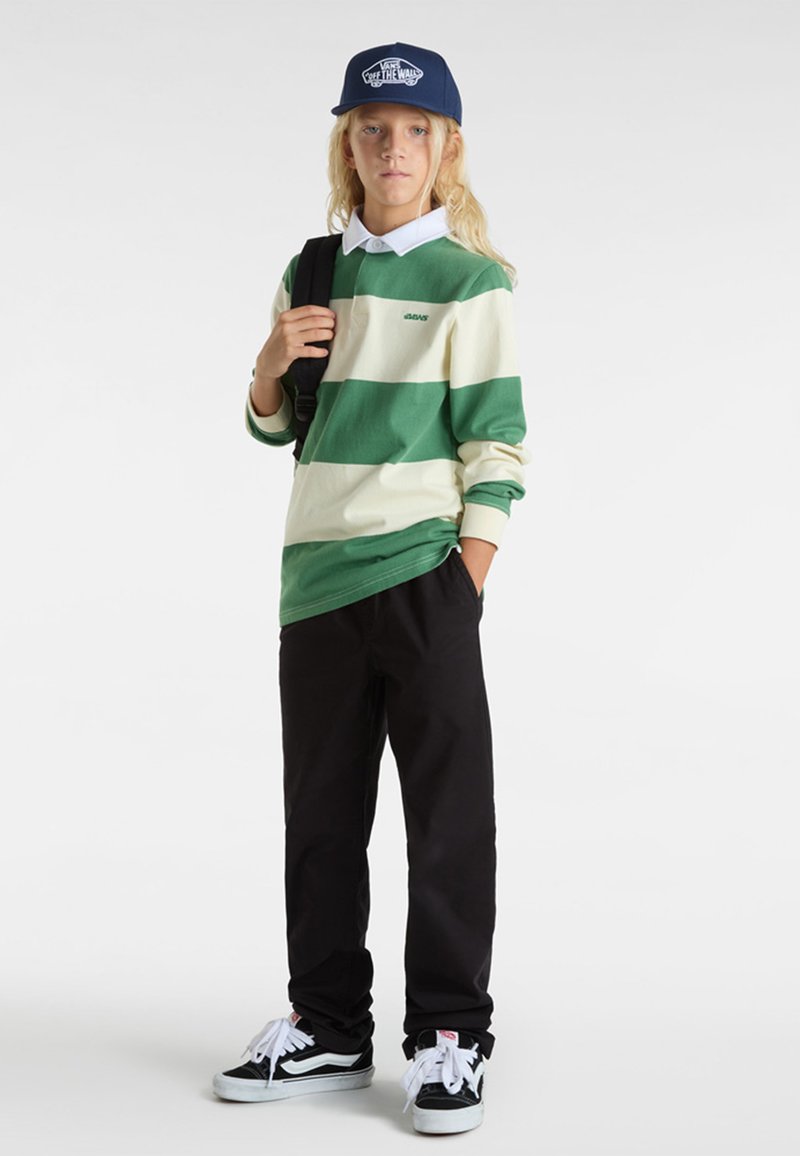 Gestreepte groene en roomkleurige lange mouwen shirt, zwarte broek, marineblauwe pet en zwarte sneakers met witte accenten, staande met een rugzak.