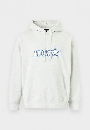 Junger Mann, der einen weißen HUF-Hoodie, schwarze grafische Jogginghosen, schwarz-weiße Sneakers und schwarze Sonnenbrillen trägt, steht vor einem weißen Hintergrund.