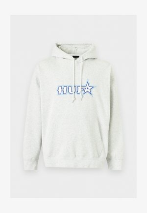 Jeune homme portant un sweat à capuche blanc HUF, un pantalon de jogging noir à motifs, des baskets noires et blanches, et des lunettes de soleil noires, debout sur un fond blanc.