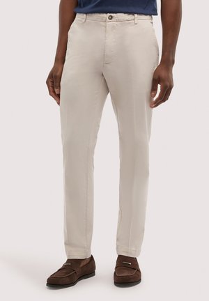 Mężczyzna ubrany w beżowe, dopasowane spodnie, ciemnobrązowe zamszowe mokasyny i granatową, włożoną do spodni koszulę, stojący na jednolitym tle.