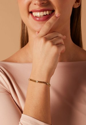 Woman smiling gently with hand near face, wearing a gold chain bracelet with small round gems, dressed in a light pink top.