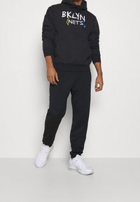 Svart hoodie med "BKLYN NETS" logotyp i vitt, gult och blått. Kombinerad med svarta joggers och vita sportskor. Slät tygtextur.